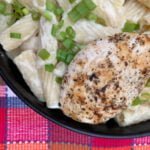 Rigatoni pasta in a white sauce topped with green onions and grilled chicken in a black bowl with a pink plaid background