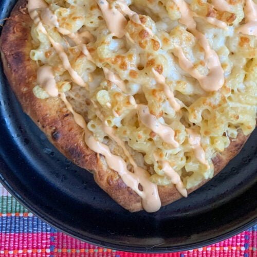 Elbow mac and cheese topped with an orange sauce on a naan crust on a black bowl with a pink plaid background