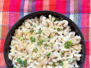 Mac and cheese in a white sauce topped with green onions and crushed chips in a black bowl with a pink plaid background