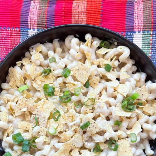 Mac and cheese in a white sauce topped with green onions and crushed chips in a black bowl with a pink plaid background