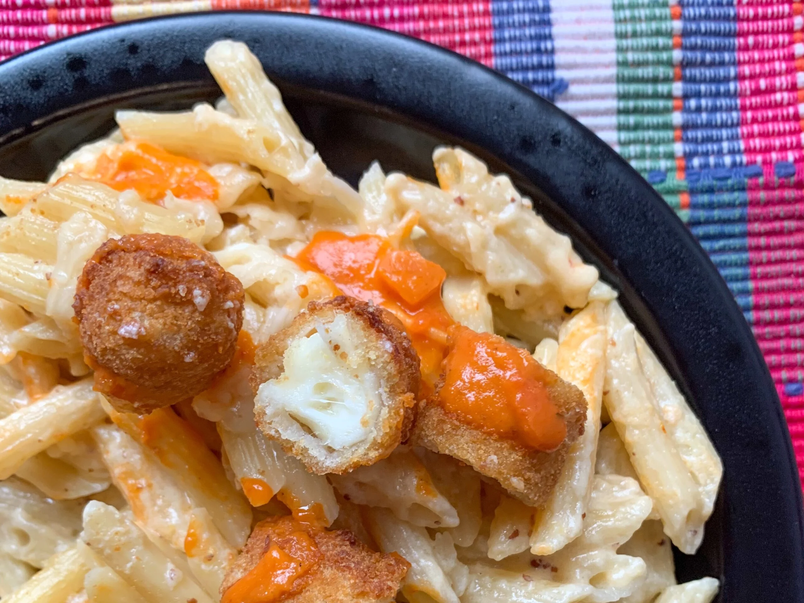 mozzarella stick mac and cheese made with Penne pasta in a white cheese sauce topped with chopped mozzarella sticks and marinara in a black bowl with a pink plaid background.